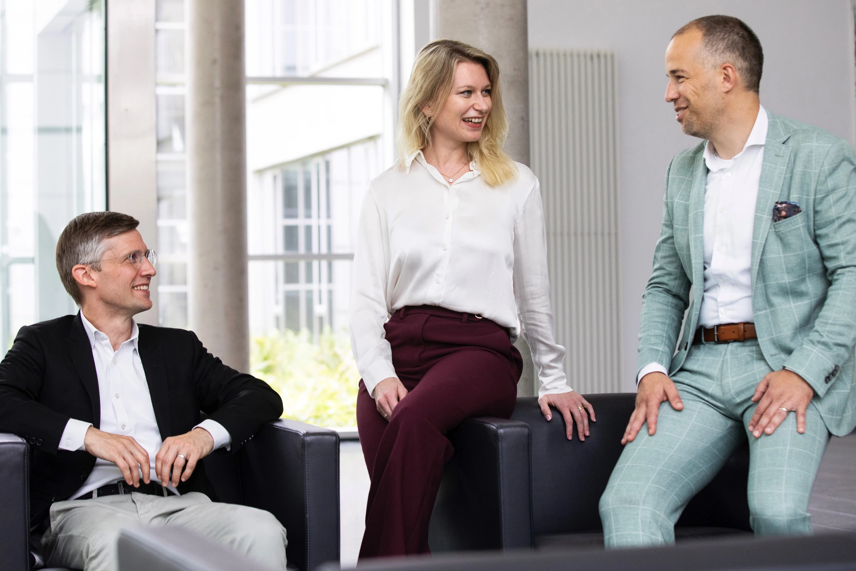 3 Geschäftsleute unterhalten sich in einem Empfangsbereich, 2 sitzen, eine Person steht – Symbolbild für lockeres Business-Meeting.