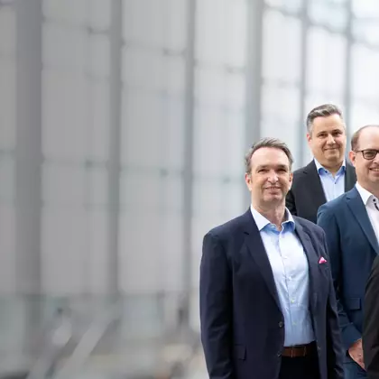 Geschäftsleute in Businesskleidung stehen vor einer modernen Glasfassade und blicken lächelnd in die Kamera – Symbolbild für Führungsteam.