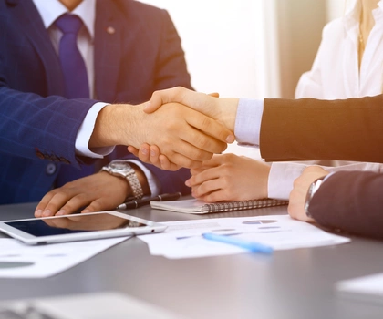 2 Geschäftspersonen geben sich die Hand über einem Schreibtisch mit Dokumenten – symbolisches Bild für erfolgreiche Zusammenarbeit.