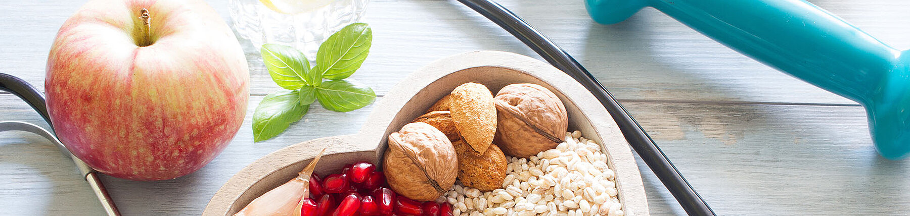 Schale mit Lebensmitteln wie Haferflocken, Nüsse und Obst, daneben ein Stethoskop und eine Hantel – Symbolbild für gesunde Ernährung und Fitness.