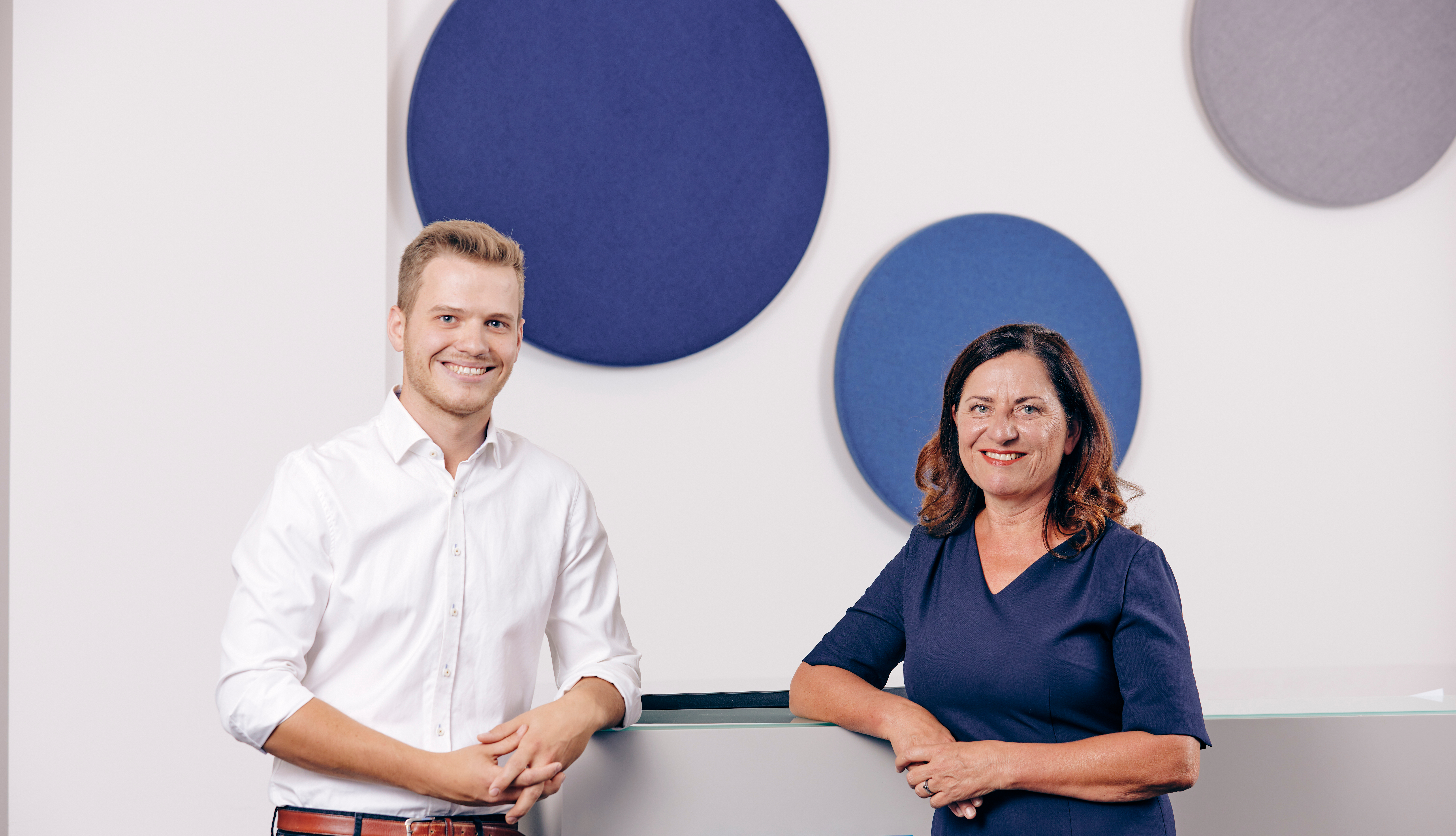 Zwei Personen stehen vor einer Wand mit dekorativen Kreisen und lächeln in die Kamera.